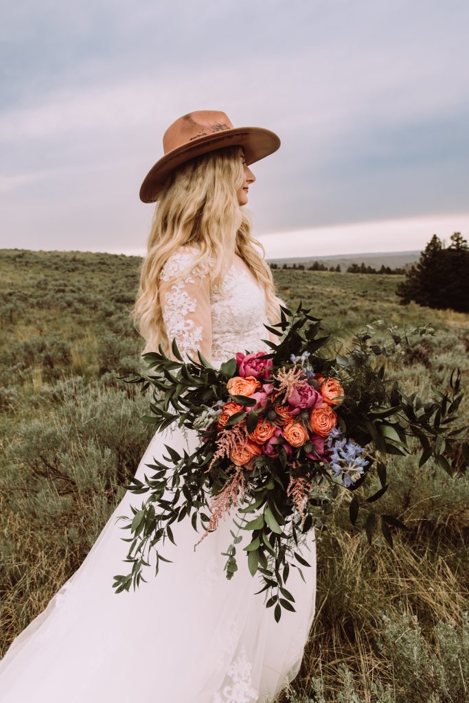 A photograph of a bride wearing a western hat carrying a bouquet with bright pink and orange flowers and dark greenery