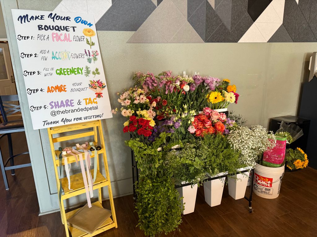 a photograph of a flower bar event setup. There is a sign on the left that says "Make Your Own Bouquet" with instructions on how to build an arrangement. On the right side are 13 buckets of florals and greenery.